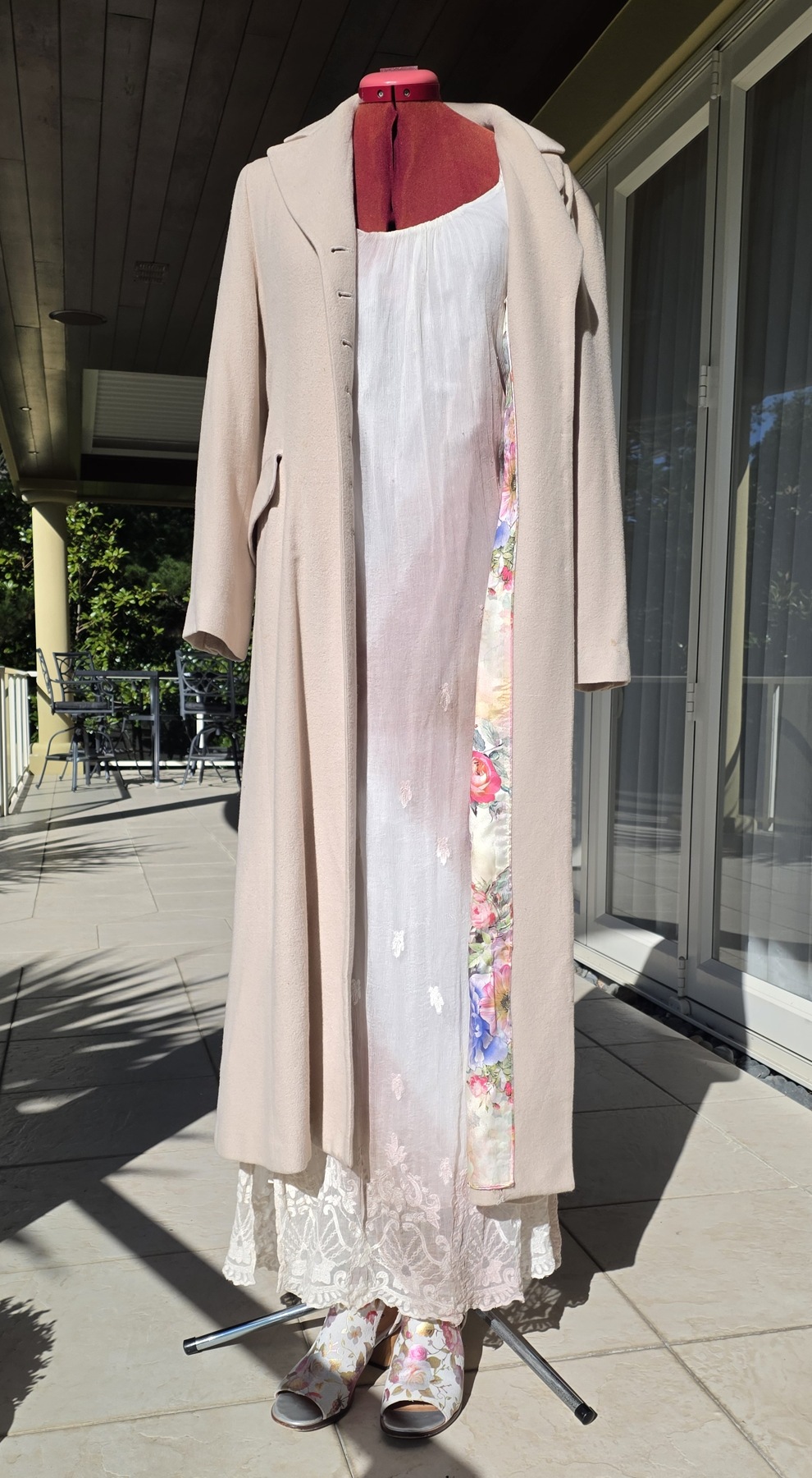 Tania’s long pale cashmere coat over a light silk chiffon dress on a dress form, with floral lining and shoes visible on a sunny terrace.
