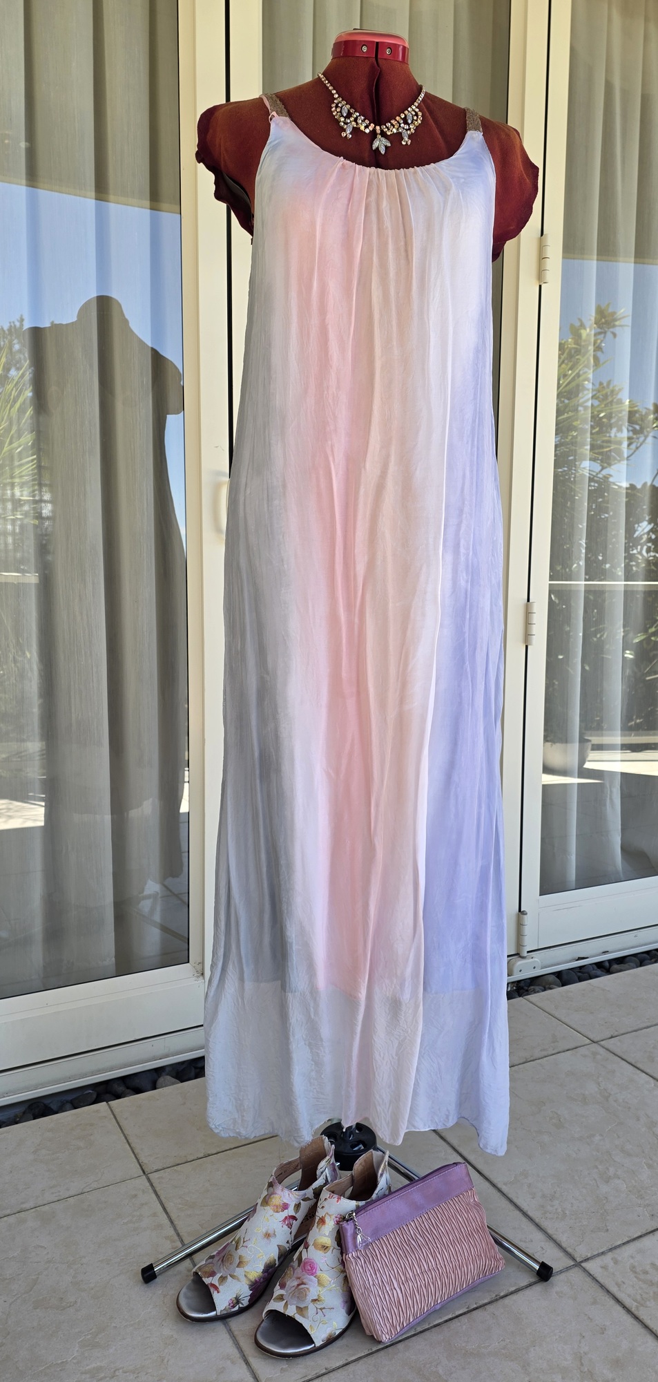 A pale long silk dress on a red mannequin with a jewelled necklace, floral shoes and a pink bag