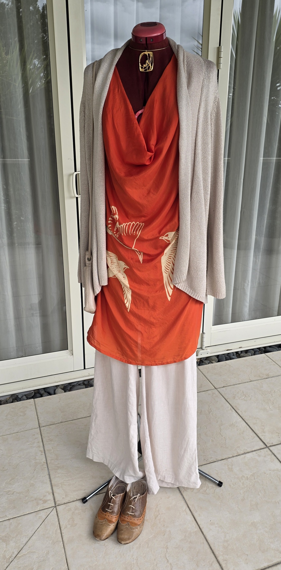 An orange silk top, pale linen trousers, a neutral longline cardigan and brown lace-up shoes on a mannequin