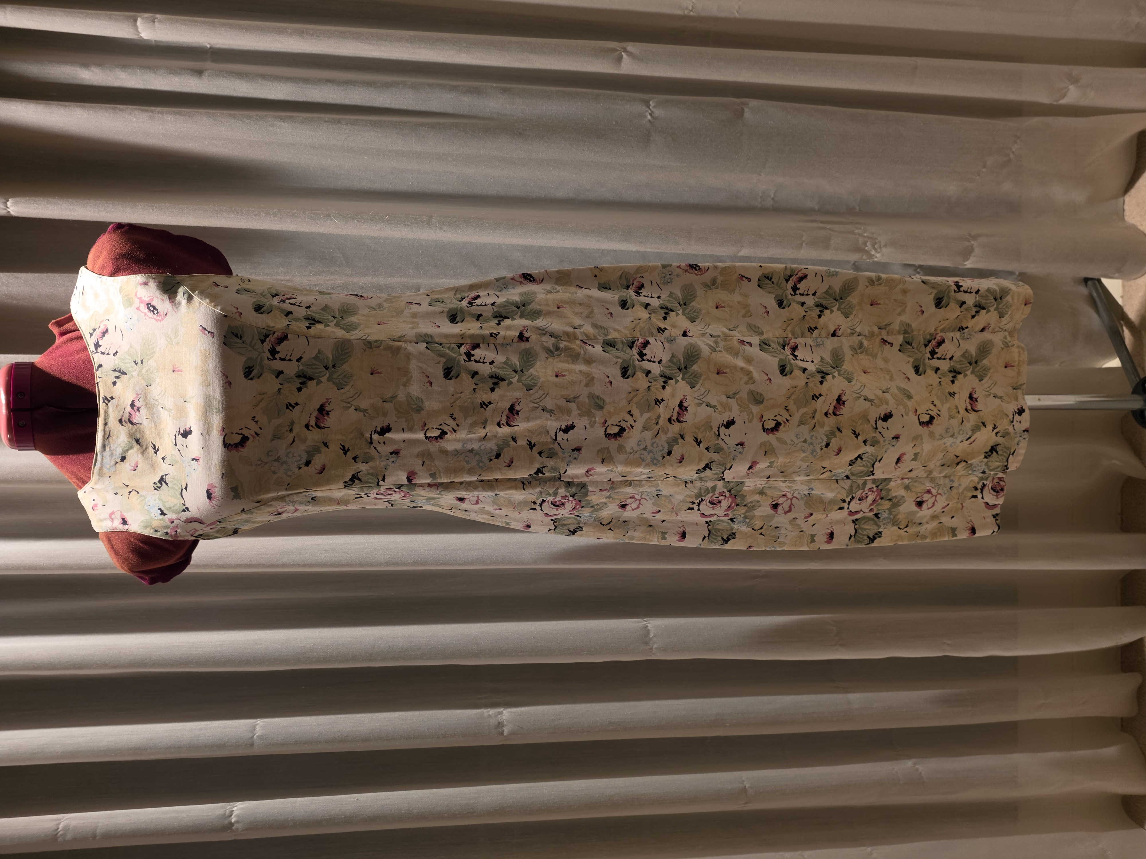 A sleeveless cream floral dress displayed on a burgundy dress form in front of pale curtains